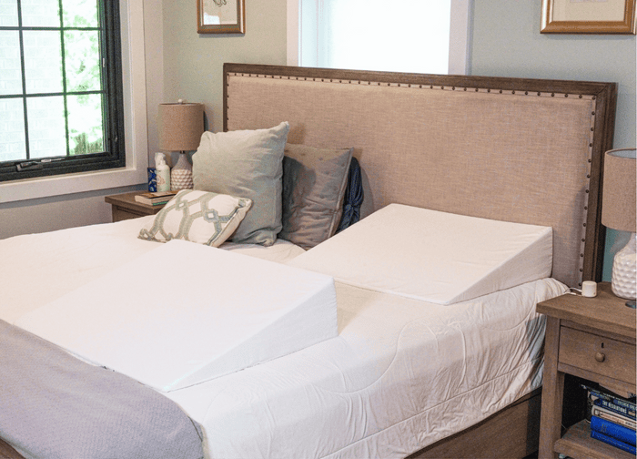 A neatly made bed with a beige headboard features two white BBL Add On Set foam wedge pillows side by side. Decorative pillows rest against the headboard, and matching lamps sit on wooden nightstands on either side.