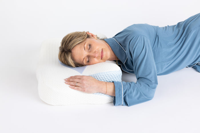 A woman in blue pajamas sleeps on her side with her head on the Cervical Head Pillow with Bamboo Cover, set against a plain white background.