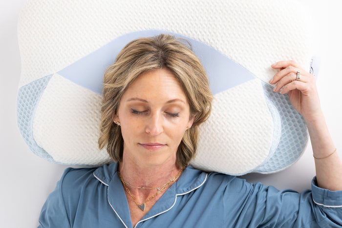 A blonde woman in blue pajamas, eyes closed, rests on the Cervical Head Pillow with Bamboo Cover. She looks relaxed and is wearing necklaces and rings.