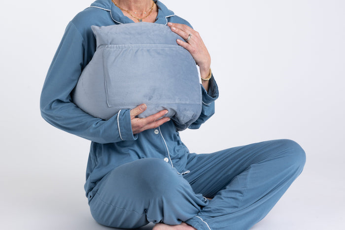A person in blue pajamas sits cross-legged, hugging the Chest Pillow—a large, light gray pillow—to their chest with both hands. The background is plain white, and the person's head is not shown.