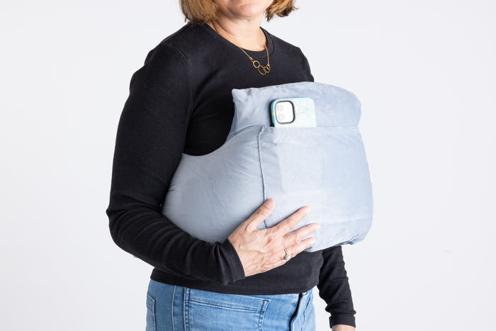 A woman in a black long-sleeve shirt and jeans holds the Chest Pillow, a large light blue post-surgery pillow with an external pocket that holds a smartphone.