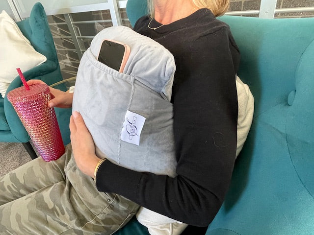 A person relaxes on a teal couch, wearing a black top and camouflage pants, using the Chest Pillow with a pocket to hold their smartphone for comfort and support after breast surgery while holding a pink tumbler.