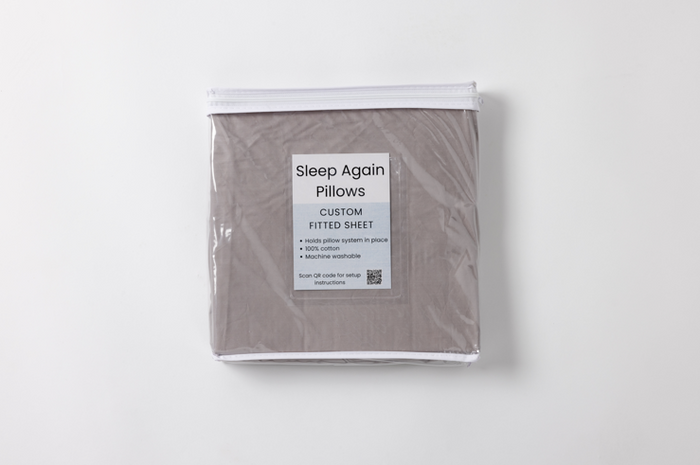 The Custom Fitted Sheet for Sleep Again Pillow System is a packaged gray, 100% cotton, machine washable sheet that helps with pillow positioning and includes a QR code for setup instructions.