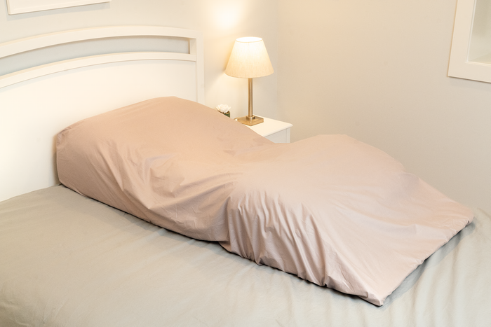 A bed features the Custom Fitted Sheet for Sleep Again Pillow System over a human-shaped pillow, resembling a person lying down; a nightstand with lamp and plant adds to the thoughtful presentation.