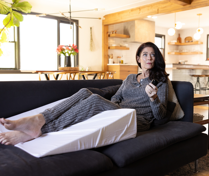 A woman in gray loungewear lounges on a modern couch, feet propped on the Individual Wedge Pillow Sold Separately, as she holds a remote and looks aside in a bright living room.