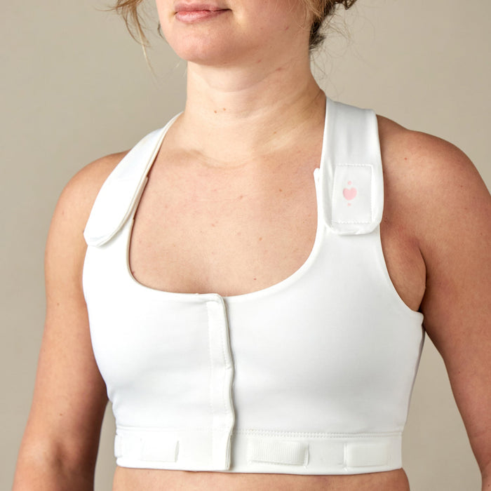 A person wears the Larissa Post-Surgical Bra, a white bra with front closures and adjustable straps for breast surgery recovery, standing against a plain beige background.