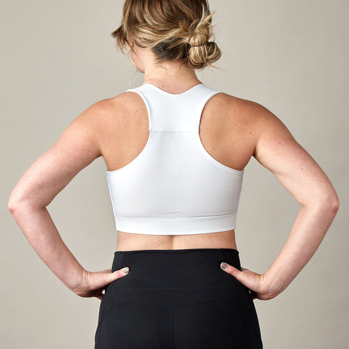 A woman with blonde hair in a messy bun stands with her hands on her hips, wearing the Larissa Post-Surgical Bra in white and black leggings against a plain background—ideal support for breast surgery recovery.