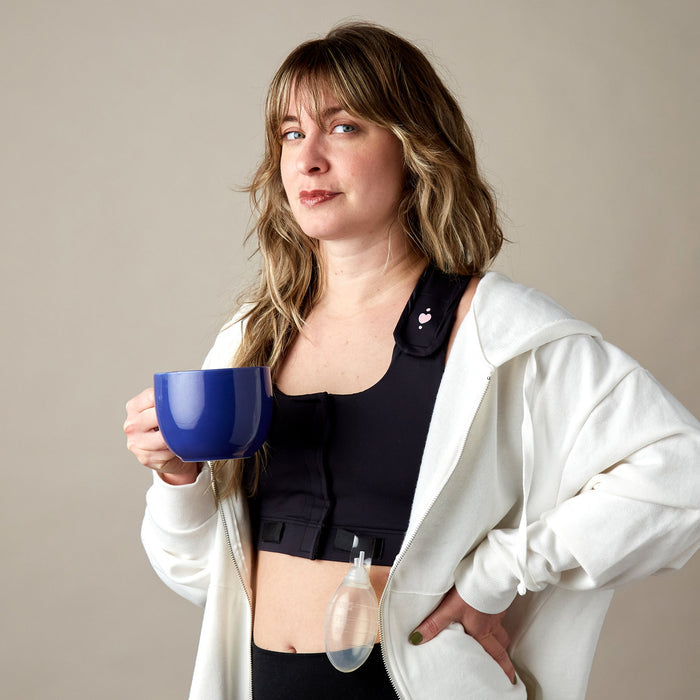 A woman with wavy hair wears a white hoodie over the Larissa Post-Surgical Bra, holding a blue mug. A surgical drain and drainage tube are attached, illustrating breast surgery recovery against a plain beige background.