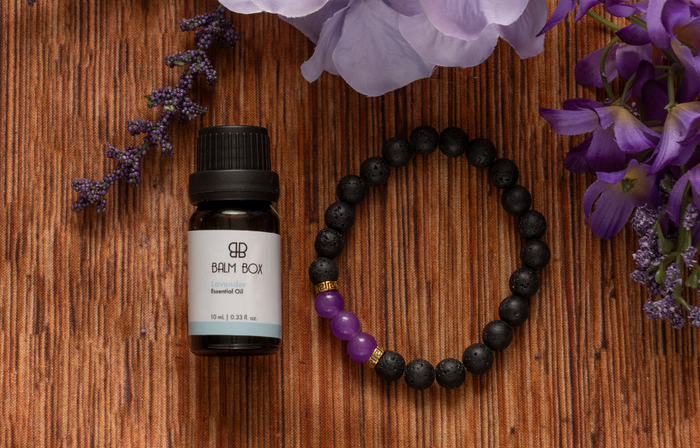 A small bottle of Lovely Lavender Essential Oil sits beside a lava-stone diffuser bracelet with purple beads on a woven surface, surrounded by purple flowers—ideal for gentle aromatherapy, especially for cancer patients.
