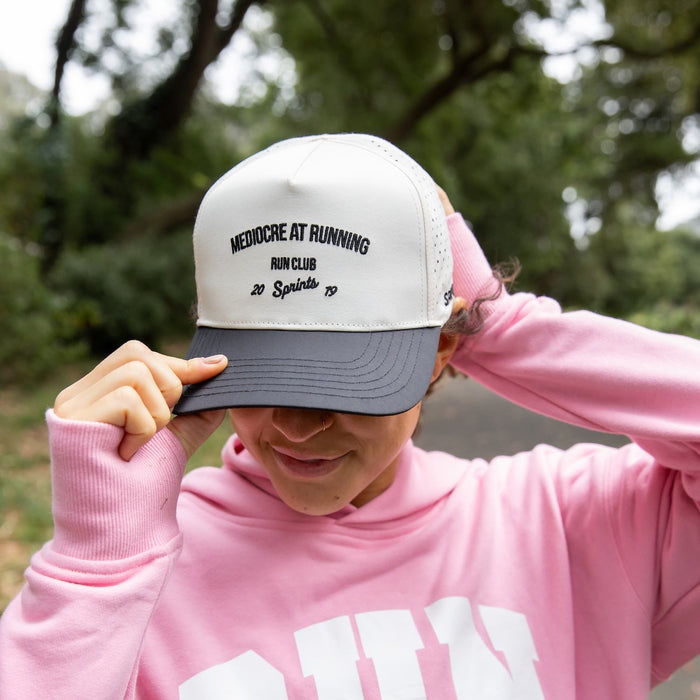 A person outdoors wearing a pink hoodie with large white 'RUN SPRINTS' text and a white-and-gray trucker cap with black embroidered text reading 'MEDIOCRE AT RUNNING RUN CLUB 20 Spirits 19'.