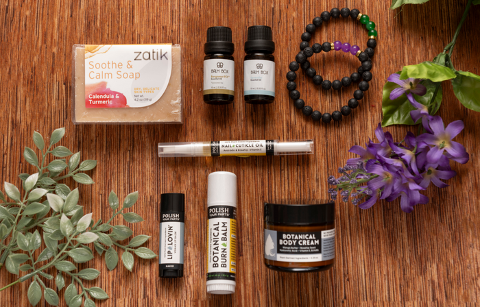 A flat lay of Premium Soothing Skin & Lips shows soap, essential oils, a cuticle oil pen, lip and burn balms, body cream, beaded bracelets, and flowers—a thoughtful self-care gift for cancer patients.