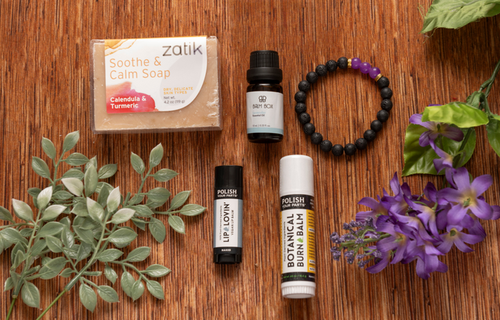Premium Soothing Skin & Lips: a flat lay set of soap, essential oil, two lip products, and a black bracelet with purple beads, accented by a purple flower—ideal for soothing care or as a thoughtful cancer support gift.