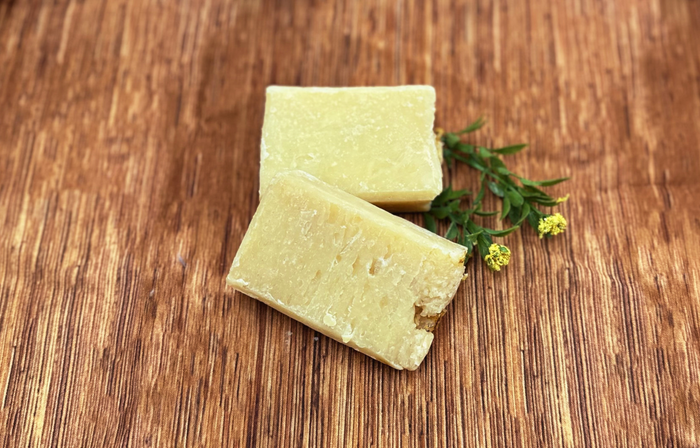 Two blocks of pale yellow Premium Soothing Skin & Lips rest on a wooden surface, adorned with a green sprig and tiny yellow flowers—an ideal addition to a soothing cancer care gift box.