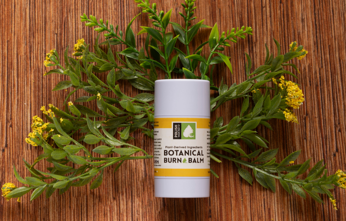 A tube of Premium Soothing Skin & Lips with a yellow label rests on a wooden surface amid green leaves and yellow flowers—a calming essential for any self-care routine, especially for cancer patients.