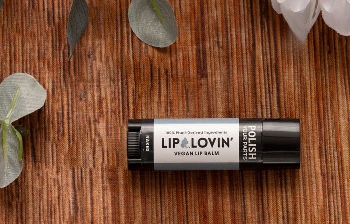 A black tube of Premium Soothing Skin & Lips balm rests on a brown textured surface amid eucalyptus and white flowers—ideal for soothing lips or adding to a cancer care gift box. The label states: “100% Plant-Derived Ingredients.”.