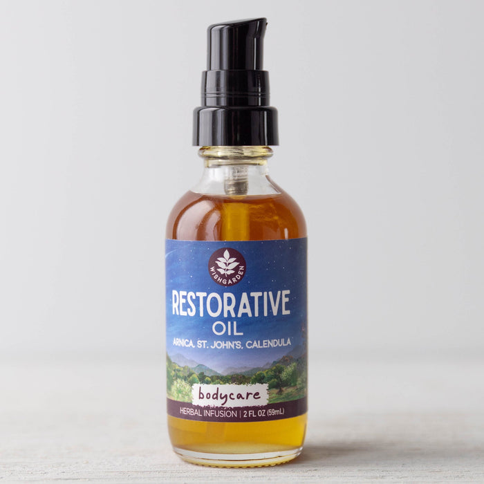 A clear glass bottle with a black pump, labeled Restorative Oil - Arnica, St. John's Wort & Calendula, sits on a light surface. The herb-infused body oil is amber in color and has a purple-blue label with white text.