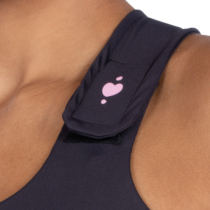 Close-up of a person’s shoulder wearing the Serena Post-Surgical Bra in a dark color, featuring a strap with a pink heart and three small pink dots. The strap is part of this reconstructive surgery garment.