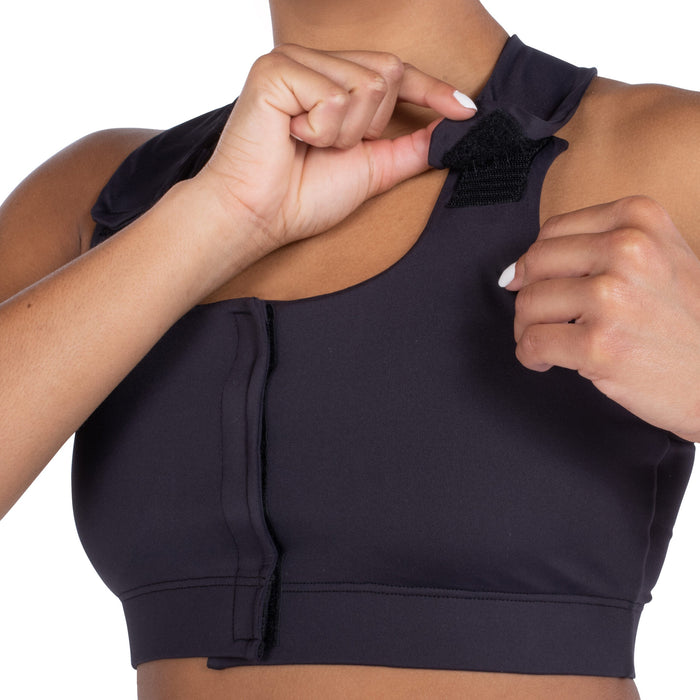 A person adjusts the Velcro strap on the shoulder of a black Serena Post-Surgical Bra, front-closing style. Only the upper torso and hands are shown against a white background.