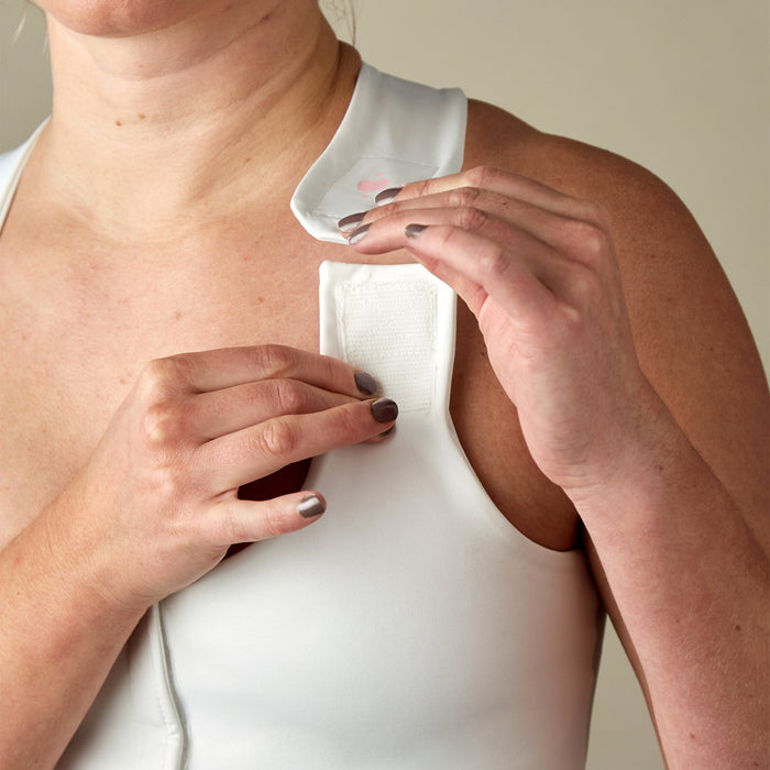 A person fastens the shoulder strap of the Serena Post-Surgical Bra, a form-fitting, sleeveless white bra with Velcro closures. Only their torso and hands are visible.