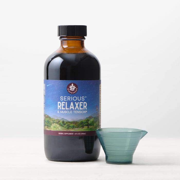 A dark glass bottle labeled "Serious Relaxer & Muscle Tension" stands next to a small, empty plastic measuring cup on a white surface, providing muscle tension relief and extra stress support.