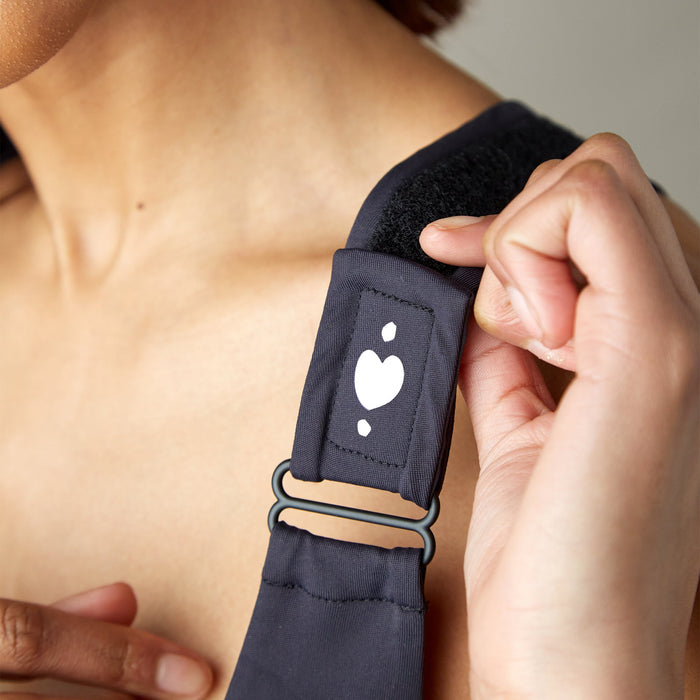 A close-up of someone adjusting the black strap of the Shirl Post-Surgical Bra on their shoulder, featuring a small white heart design and a hand holding the strap near its metal buckle.