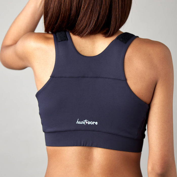 A woman with shoulder-length brown hair stands against a plain light background, wearing the Shirl Post-Surgical Bra in dark color with "heartcore" printed in white on the lower back.