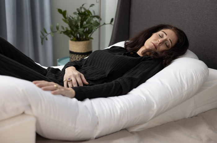 A woman with dark hair lies on a bed, eyes closed, hugging the Sleep Again Pillow System—a pillow designed for side sleepers. A potted plant is visible in the background.