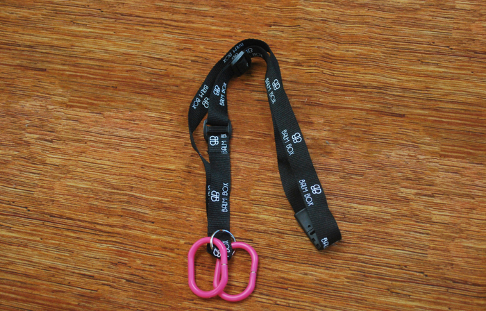 A black “BHA BOX” Surgical Drain Holder lanyard with white text and logo rests on a wooden surface. It features two connected pink plastic clips at one end, making it ideal for use as a mastectomy drain holder.
