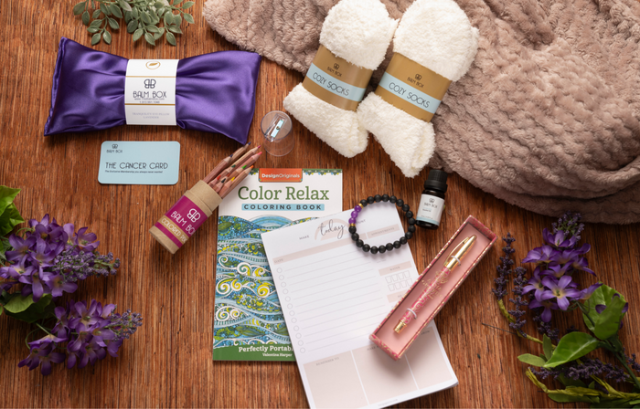 The Sweetest Rest & Recovery Box is a cozy cancer recovery gift featuring a purple sleep mask, fuzzy socks, bath soak, planner, coloring book, pen set, bracelet, essential oil, gift card, and purple flowers on a soft brown blanket.