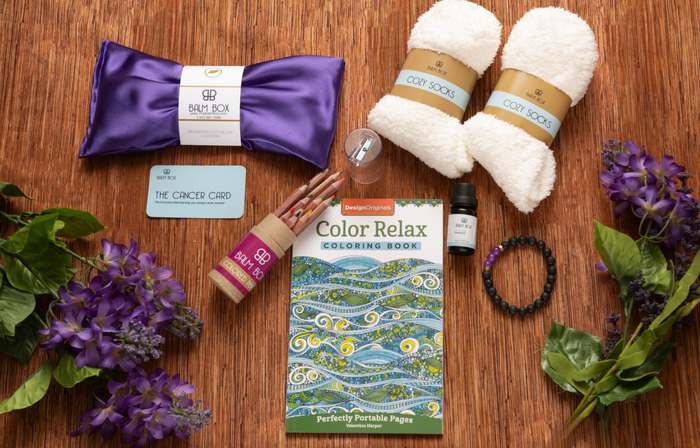 The Sweetest Rest & Recovery Box on a wooden surface includes cozy socks, a lavender eye pillow, an inspirational coloring book, colored pencils, essential oil, a bracelet, lip balm, a Cancer Card, and purple flowers.