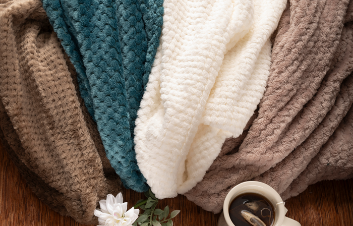 Four soft, textured blankets in brown, teal, white, and taupe are draped beside a white mug of tea and flower—offering cozy comfort found in the Sweetest Rest & Recovery Box, perfect for any cancer recovery gift.
