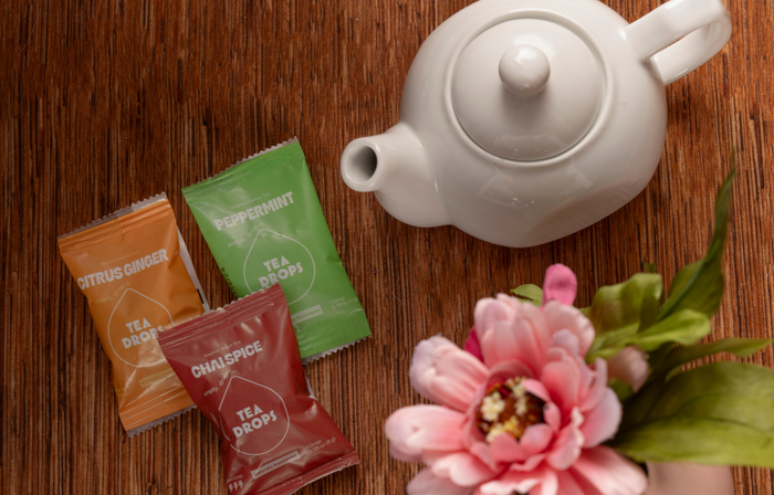 A white teapot sits on wood next to Tea Drops in citrus ginger, peppermint, and chai spice from the Sweetest Rest & Recovery Box—ideal for self-care or cancer recovery. A pink artificial flower with green leaves adds a soft finishing touch.