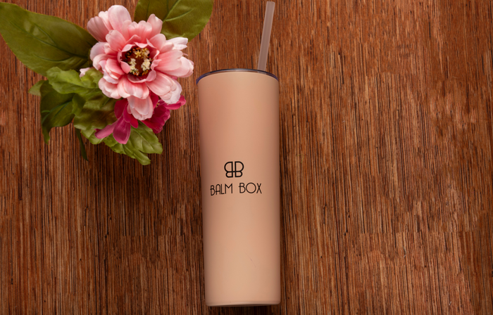 A beige tumbler with straw and BALM BOX logo sits on a wooden surface beside a pink flower—an ideal addition to the Sweetest Rest & Recovery Box, a thoughtful self-care gift for cancer patients.