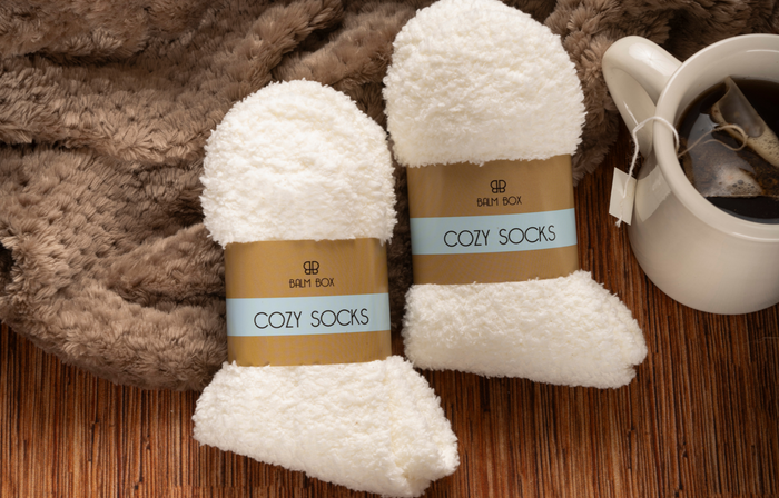 Two pairs of fluffy white Cozy Socks sit on a brown textured surface beside a soft blanket and a white mug—an ideal addition to the Sweetest Rest & Recovery Box or perfect for self care for cancer patients.