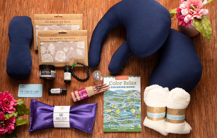 A flat lay of The Best Mastectomy Care Package features a neck pillow, ice packs, cozy socks, colored pencils, coloring book, satin eye mask, flowers, bracelet, candle, and balm—thoughtfully assembled for breast cancer or mastectomy recovery.