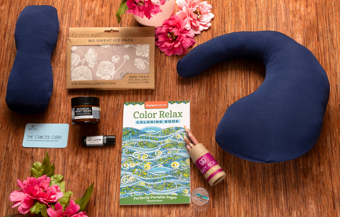 The Best Mastectomy Care Package on a wooden surface features a blue neck pillow, eye mask, coloring book, lip balm, lotion, chill pack, gift card, and pink flowers—ideal for comfort and support after a mastectomy.