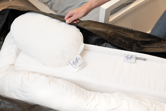 A hand lifts the black Travel and Storage Bag for Sleep Again Pillow System, revealing a white body pillow with tags inside, water resistant and ideal for travel, next to a bed with gray sheets in the background.