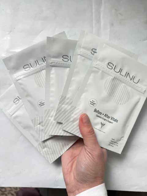 A hand displays five white SULINU Travel Packs—Set of 5, Single Serving Before + After Vitals—vitamin powder, fanned out against a light background. Designed to aid wound healing after surgery and support immunity.
