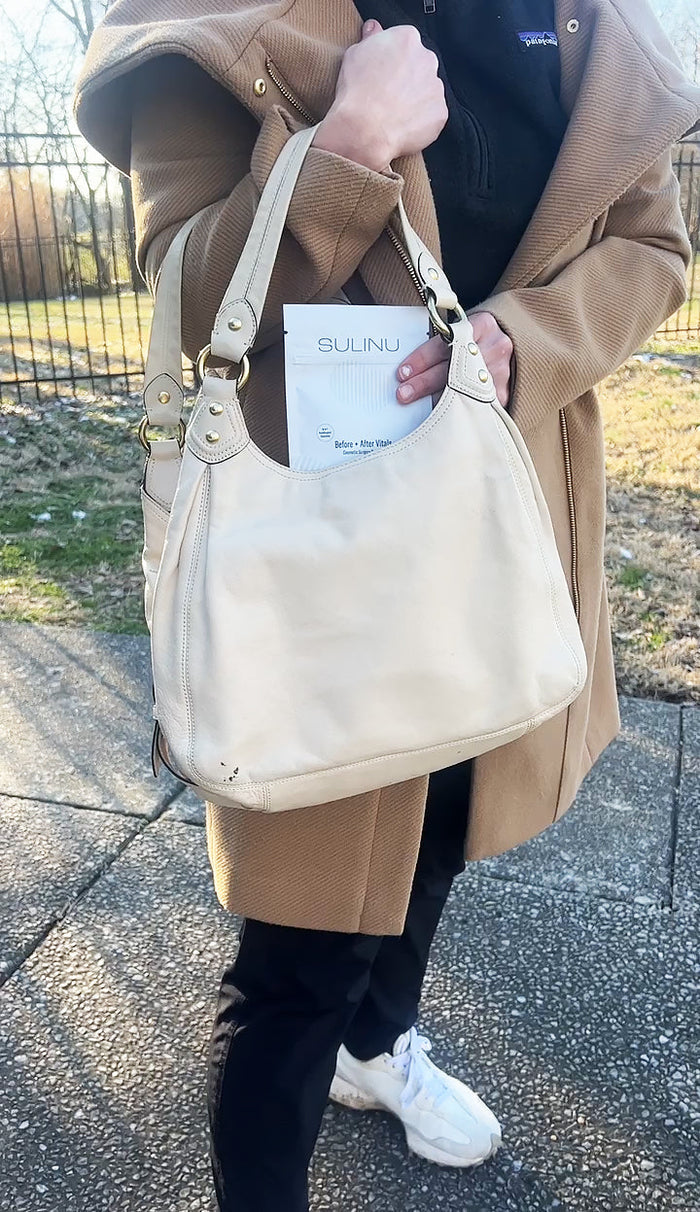 A person in a tan coat and black pants holds a beige handbag with a document labeled SULINU partly visible, possibly carrying Travel Packs- Set of 5- Single Serving Before + After Vitals to support wound healing after surgery.
