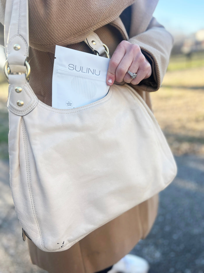 A person in a tan coat places a white Travel Packs-Set of 5-Single Serving Before + After Vitals vitamin powder packet into a light beige purse’s front pocket outdoors, with blurred greenery and pavement in the background.